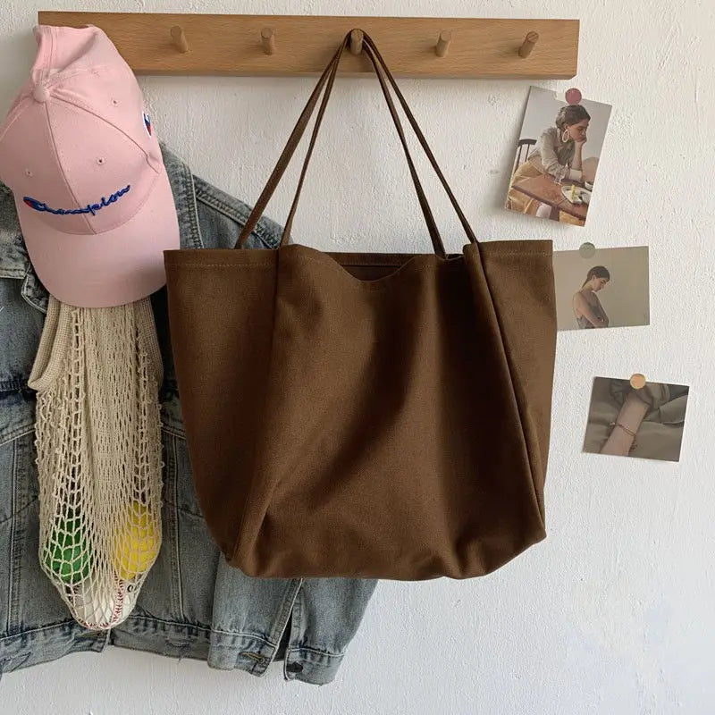 Brown tote bag hanging on a wooden hook with a pink cap, denim jacket, and mesh bag below.