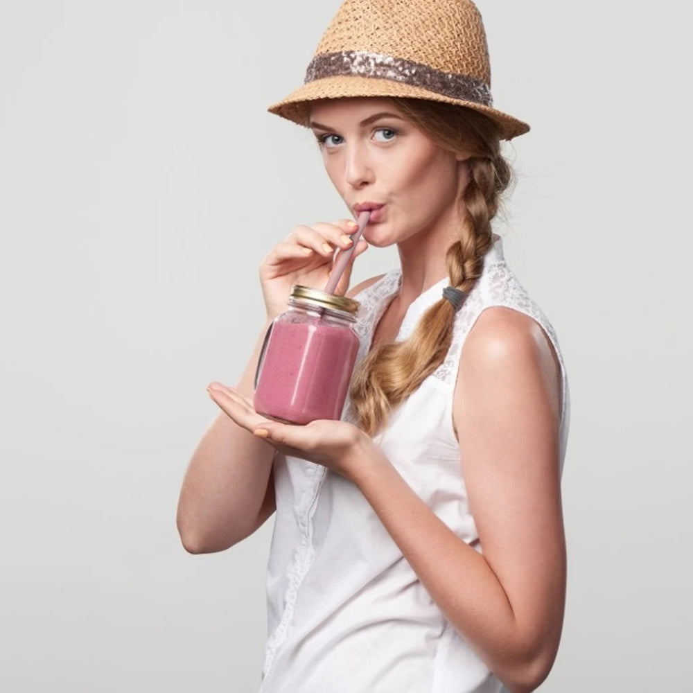 Woman drinking a pink smoothie from a jar with a straw, wearing a straw hat.