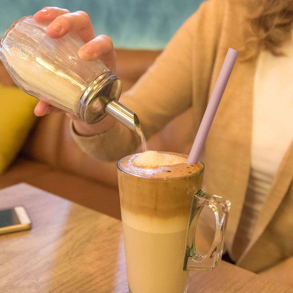 Person pouring a liquid into a glass of coffee with a straw on a wooden table.